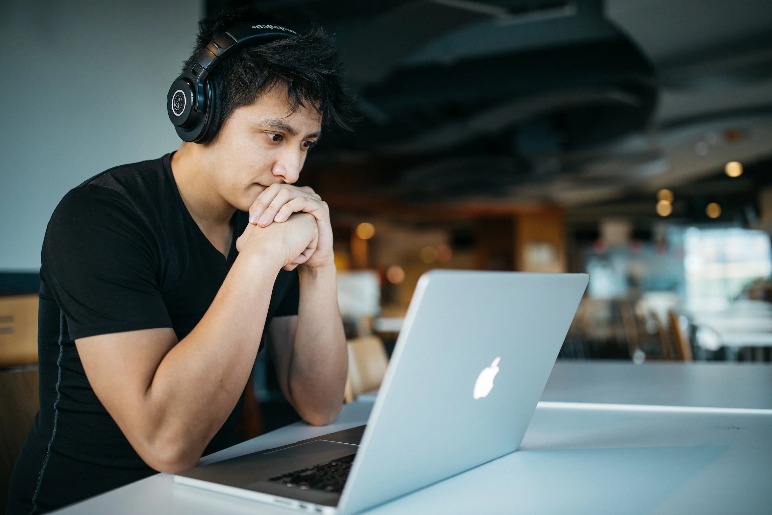 A young man with black hair wearing headphones, sitting at a table in a modern cafe, looking intently at a silver MacBook laptop.